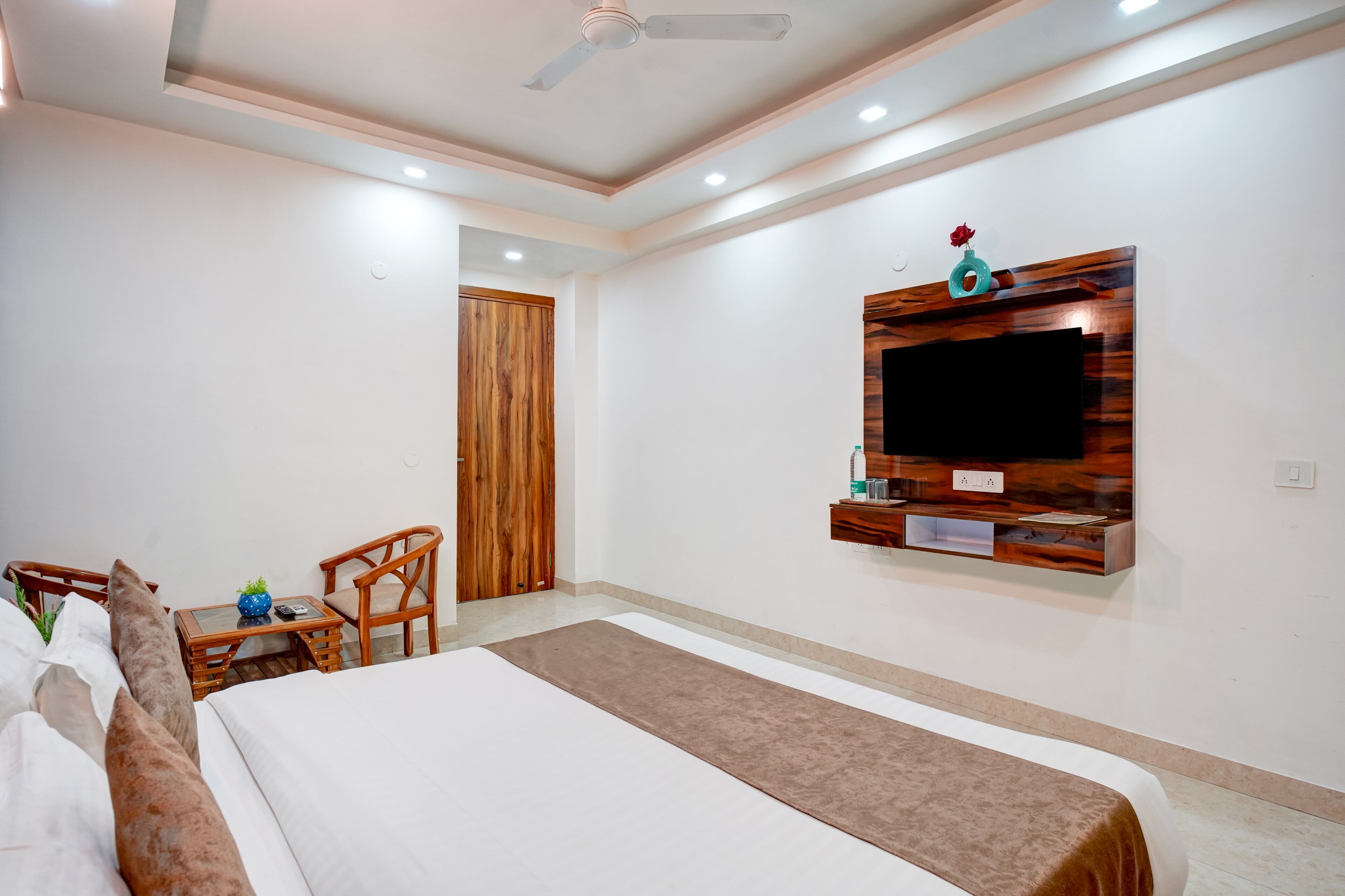 A hotel room with a bed, a TV mounted on the wall, a small table with chairs, and a wooden door.