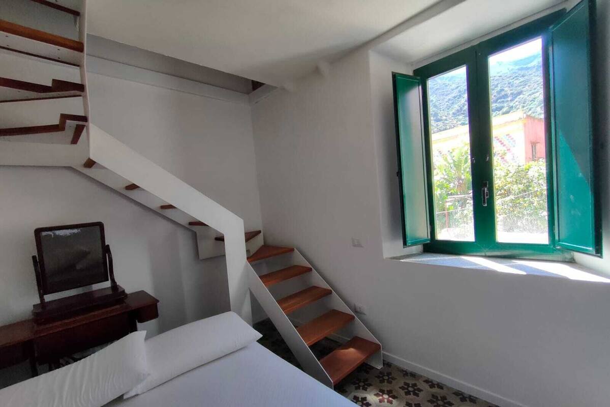 A bedroom with a staircase, a bed, a TV, and a window with green shutters.