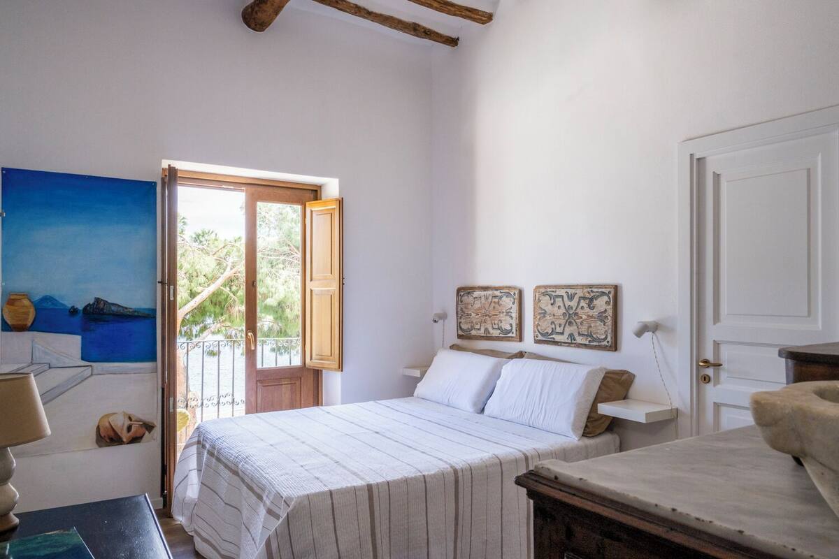A bedroom with a bed, a door, a window, and a painting on the wall.