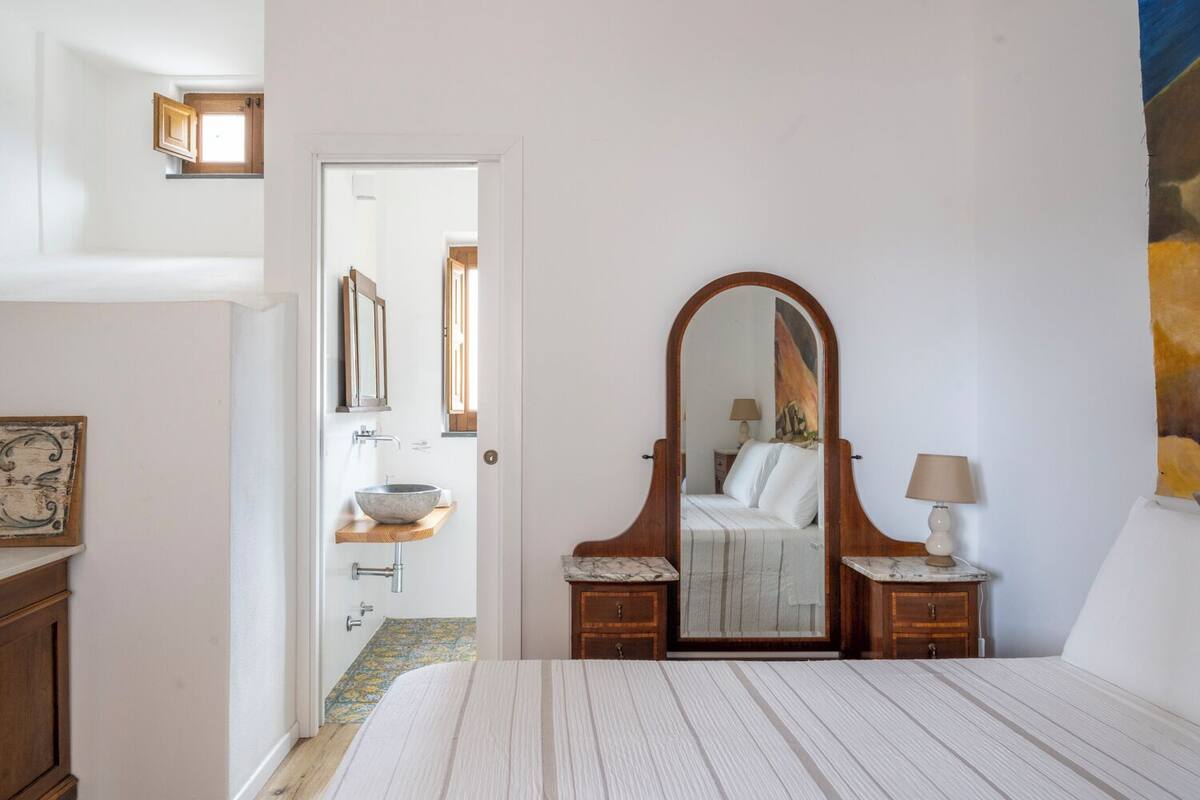 A bedroom with a bed, a wooden bedside table with a lamp, a large mirror, and a bathroom with a sink and cabinet.
