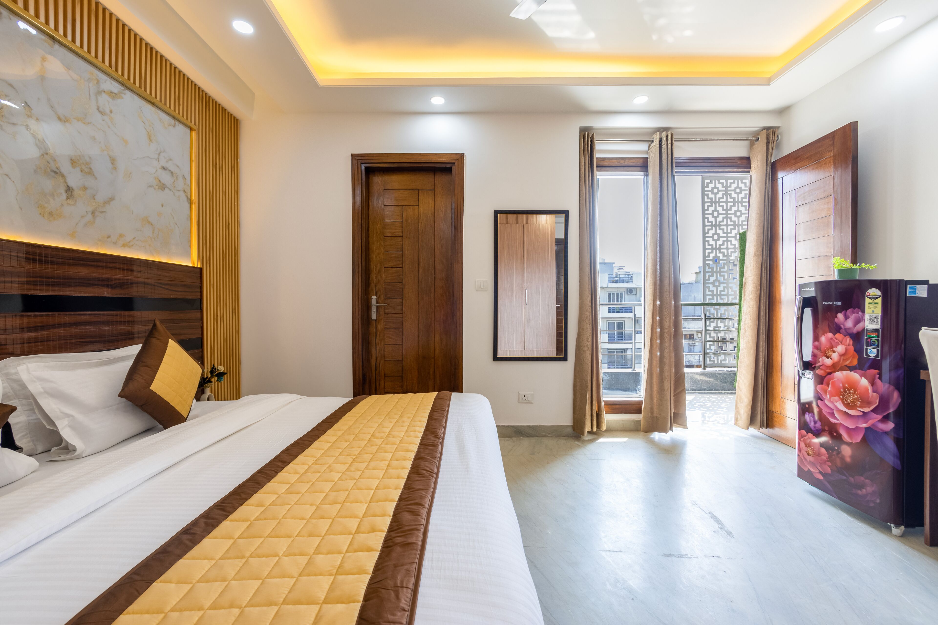 A hotel room with a bed, wooden headboard, a large mirror, a refrigerator with floral design, and a view of the outside.