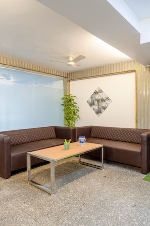 Uma área de espera moderna com um sofá de couro marrom, uma mesa de metal com uma planta e garrafas de água, um ventilador de teto e uma parede com uma obra de arte geométrica.