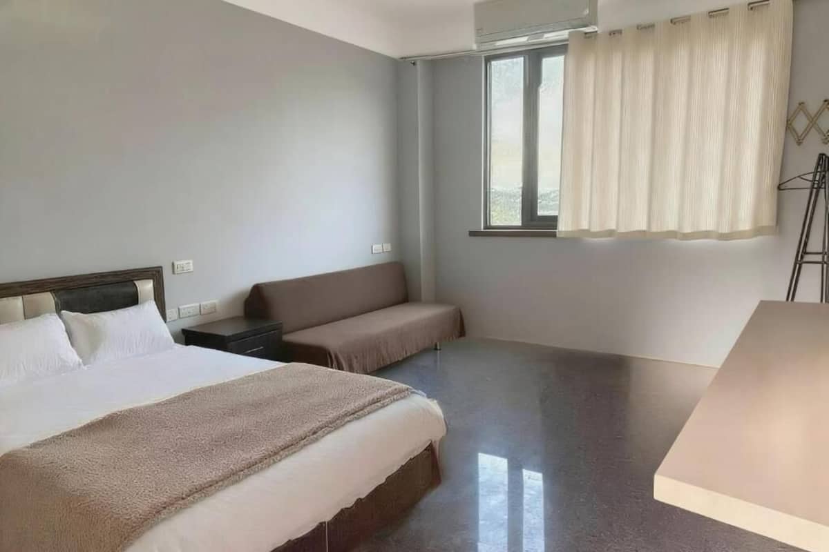 Habitación de hotel con una cama grande, un banco, una mesita de noche, una ventana con cortinas y una lámpara de pie.