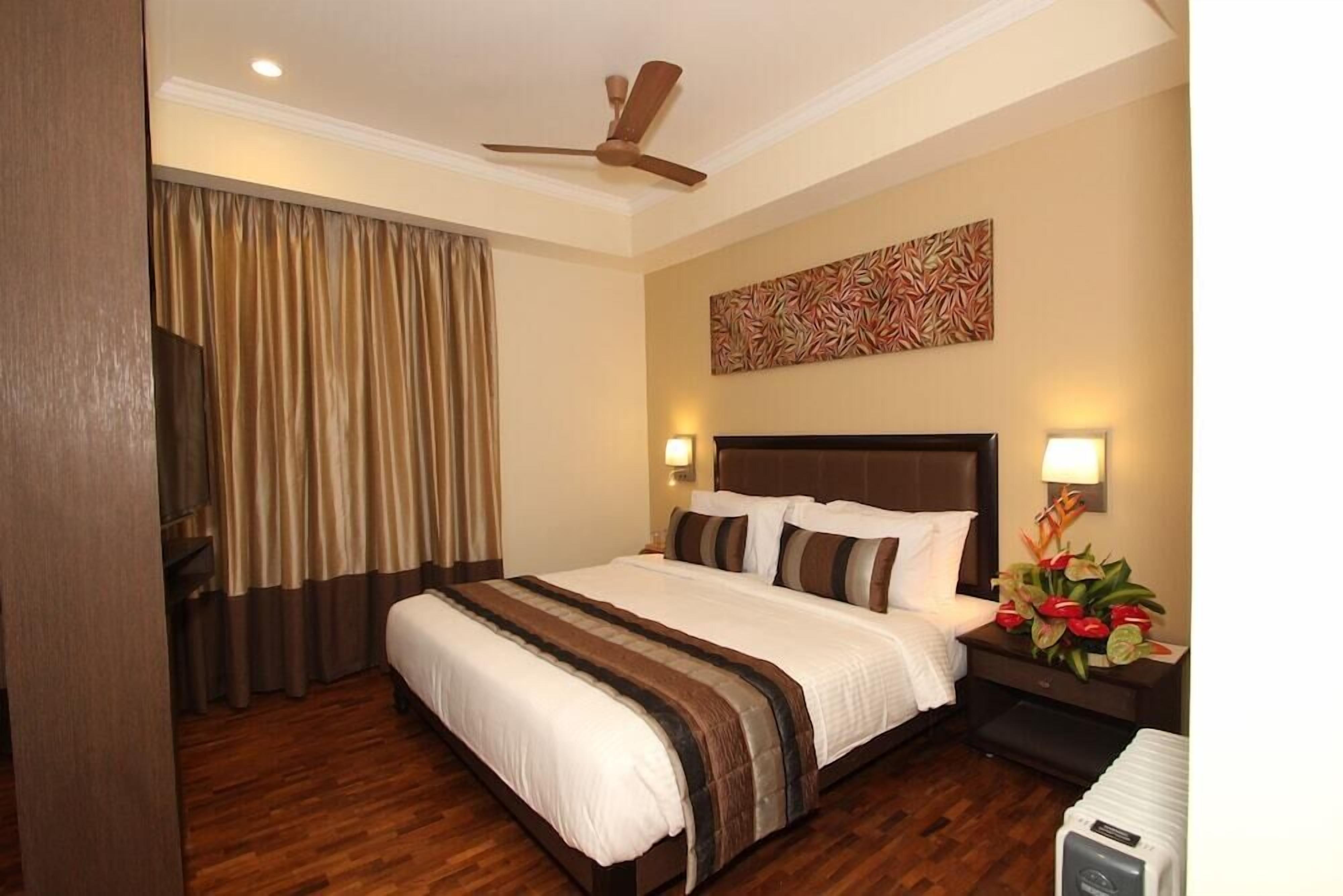 A hotel room with a large bed, wooden flooring, a ceiling fan, a wall-mounted TV, and a nightstand with a lamp and flowers.