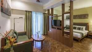 A hotel room with a sofa, a small table with a vase of flowers, a bed with a TV, and a wooden headboard. - Club Mahindra Mount Serene Munnar (Devikolam)