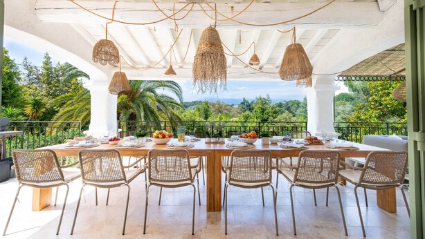 An outdoor dining area with a table set for six, surrounded by wicker chairs, under a white pergola with hanging lights, overlooking a lush garden and distant mountains. - Villa Anna on the Côte d'Azur | 8 bedrooms, private Padel and P (Valbonne)