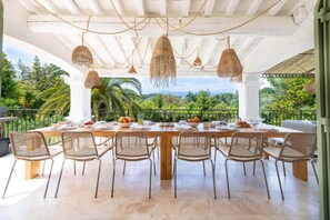 Un espace de restauration en plein air avec une table dressée pour six personnes, entourée de chaises en osier, sous une pergola blanche agrémentée de guirlandes lumineuses, offrant une vue sur un jardin luxuriant et des montagnes lointaines.