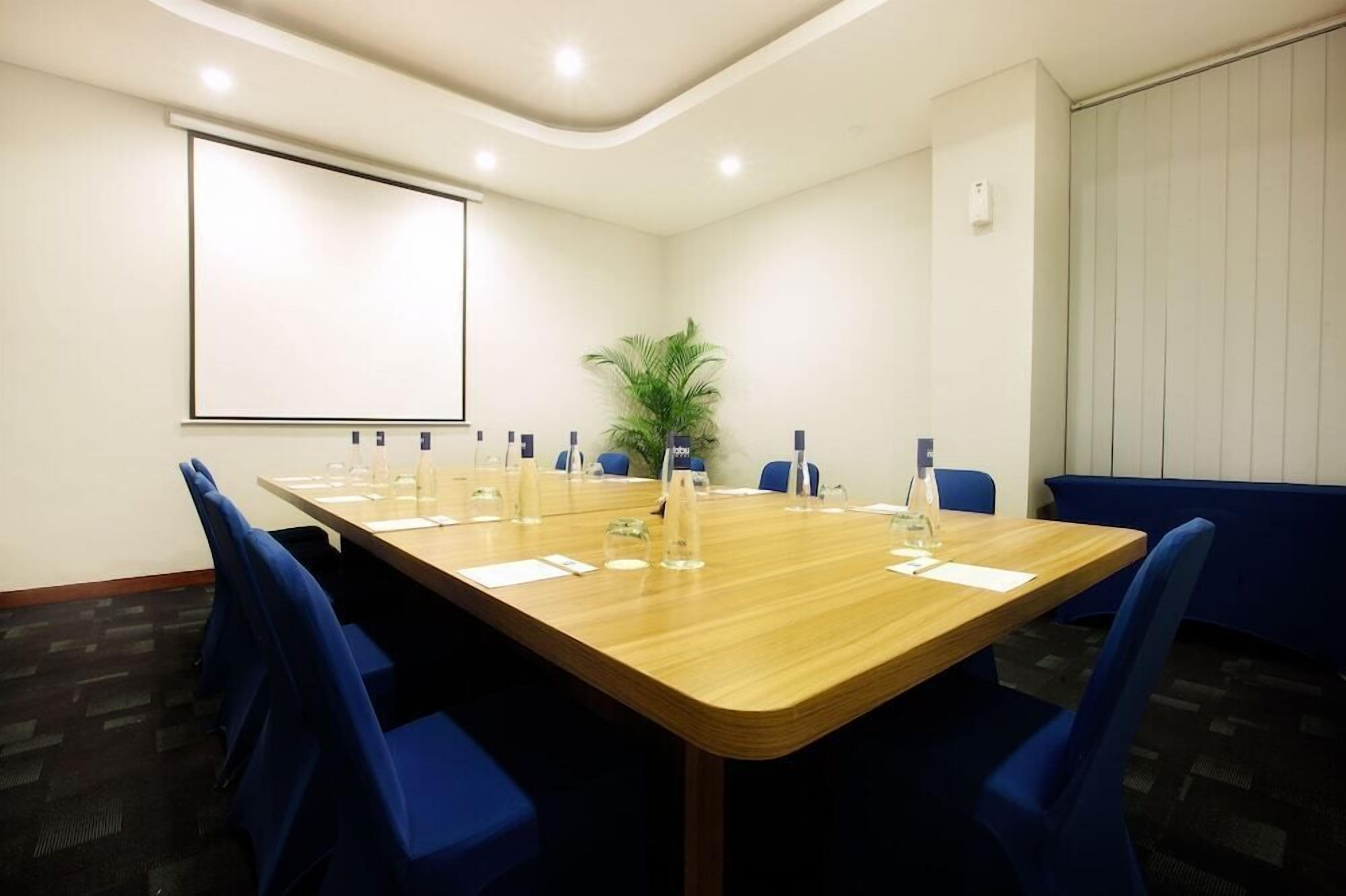 Une salle de conférence avec une longue table, des chaises bleues, un écran de projection et une plante en pot.
