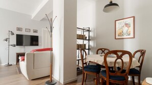 Ein Wohnzimmer mit einer Couch, einem Esstisch mit Stühlen, einem Bücherregal, einem Fernseher und einer Lampe.