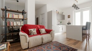 Ein Wohnzimmer mit einer Couch, einem gemusterten Teppich, einem Bücherregal und einer Küchenzeile im Hintergrund.
