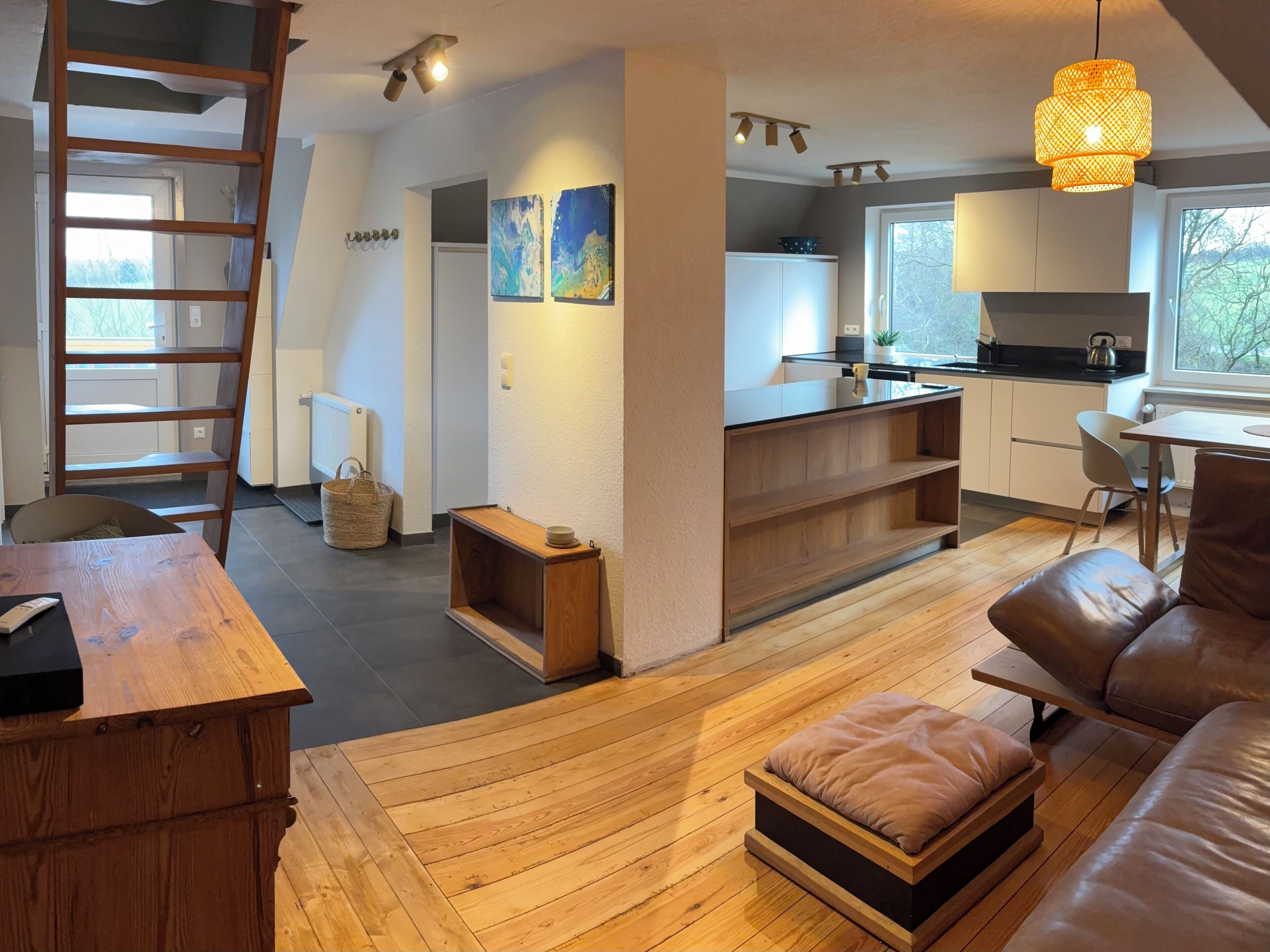 A modern living room with a wooden floor, a brown leather sofa, a wooden coffee table, and a wooden side table. There is a kitchen area with white cabinets and a black countertop. A ladder is leaning against the wall, and there are two paintings on the wall.