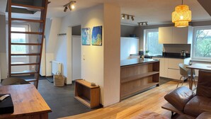 Een moderne woonkamer met een houten vloer, een bruine leren bank, een houten salontafel en een houten bijzettafeltje. Er is een keuken met witte kasten en een zwart aanrecht. Een ladder staat tegen de muur, en er hangen twee schilderijen.