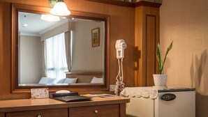 Habitación de hotel con tocador de madera, espejo, secador de pelo, planta en maceta y refrigerador antiguo.