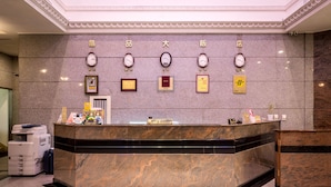 Zona de recepción de hotel con un mostrador de mármol, relojes mostrando diferentes husos horarios, certificados enmarcados y una caja registradora.