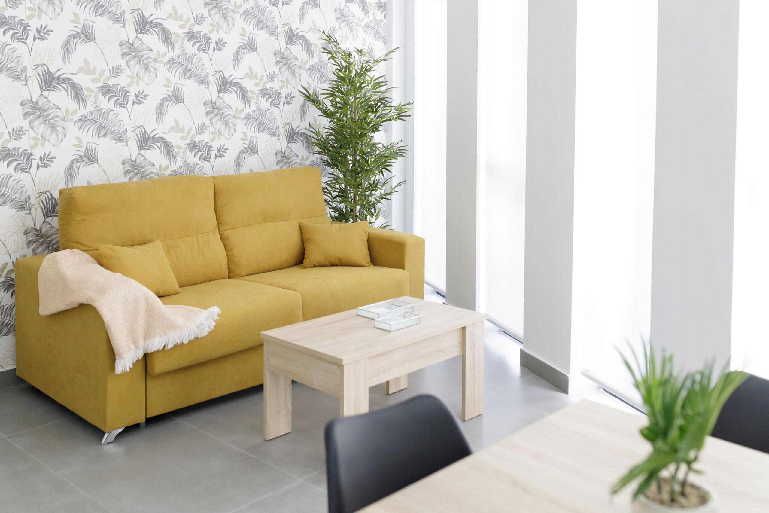Uma sala de estar moderna com um sofá amarelo mostarda, uma mesa de centro de madeira e uma grande janela com vista para o exterior.
