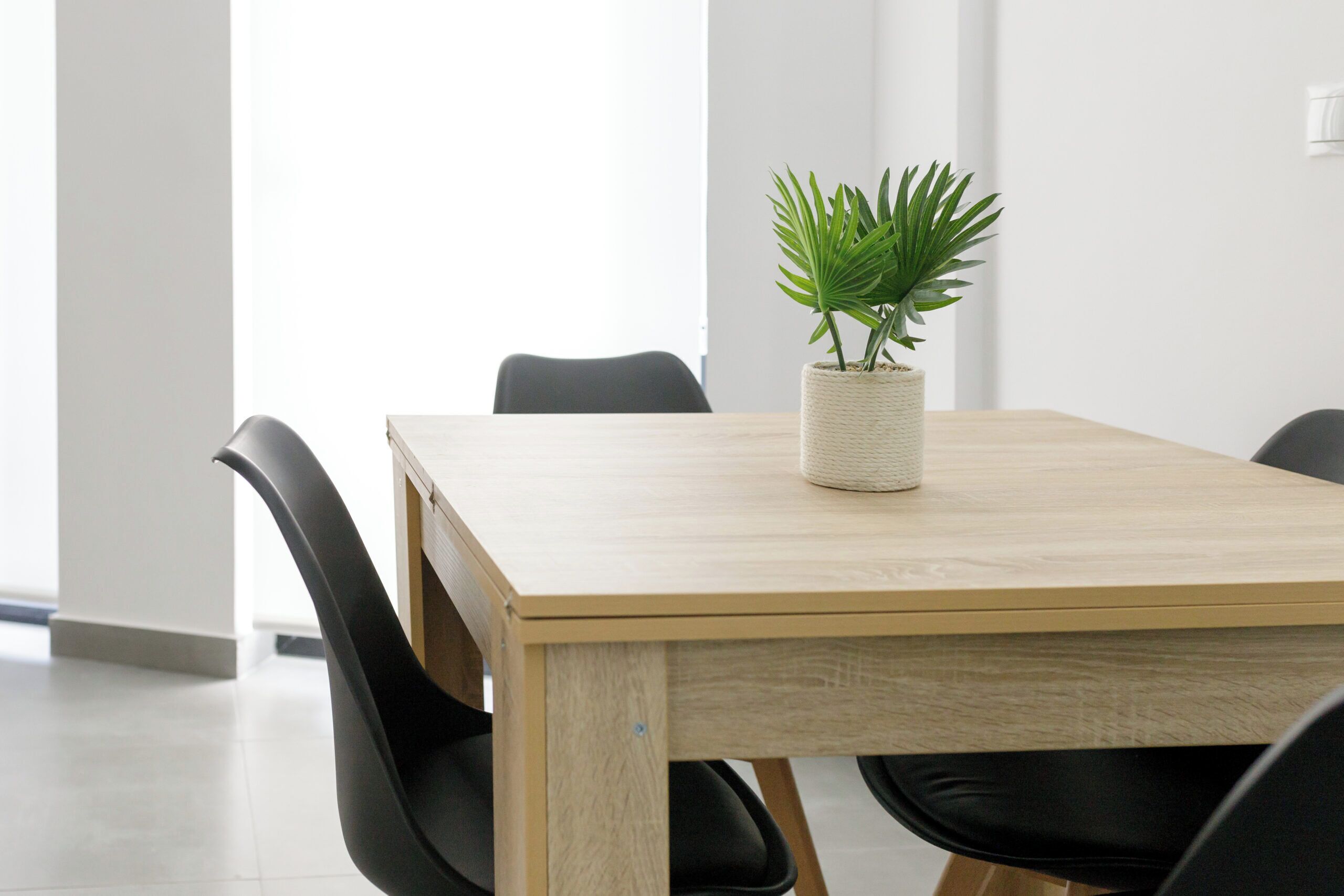 Una mesa de madera con una maceta tejida, sillas negras y una pared blanca al fondo.