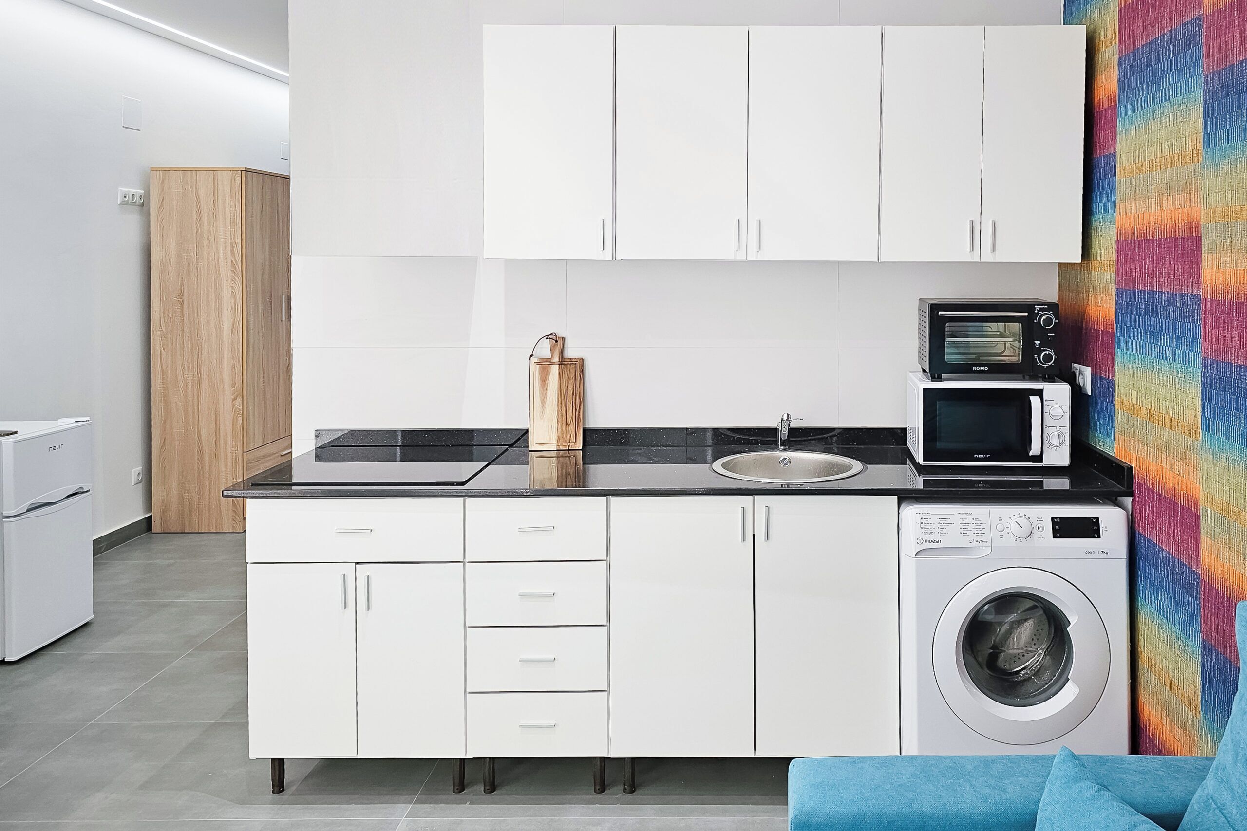 Une cuisine moderne avec des meubles blancs, un four à micro-ondes, un lave-linge et un plan de travail noir.