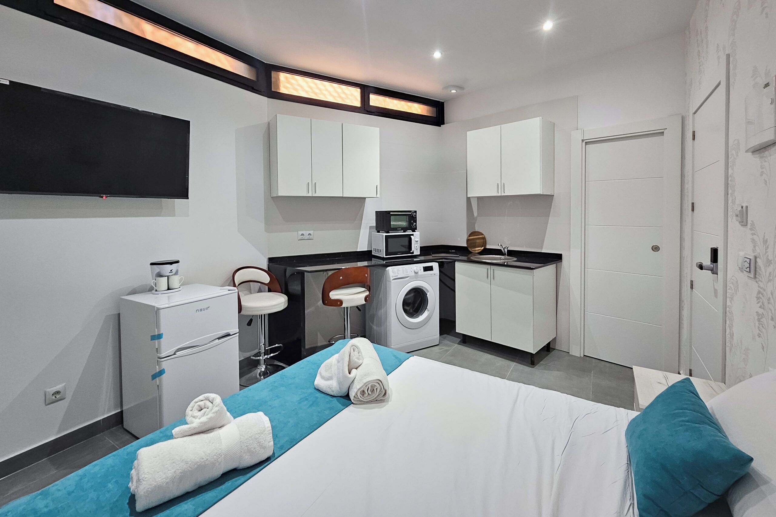Une chambre d’hôtel compacte comprenant un lit, une petite kitchenette, un lave-linge et un téléviseur à écran plat.