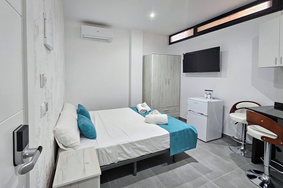 A modern hotel room with a bed, a desk, a chair, a wardrobe, a TV, and a minibar.