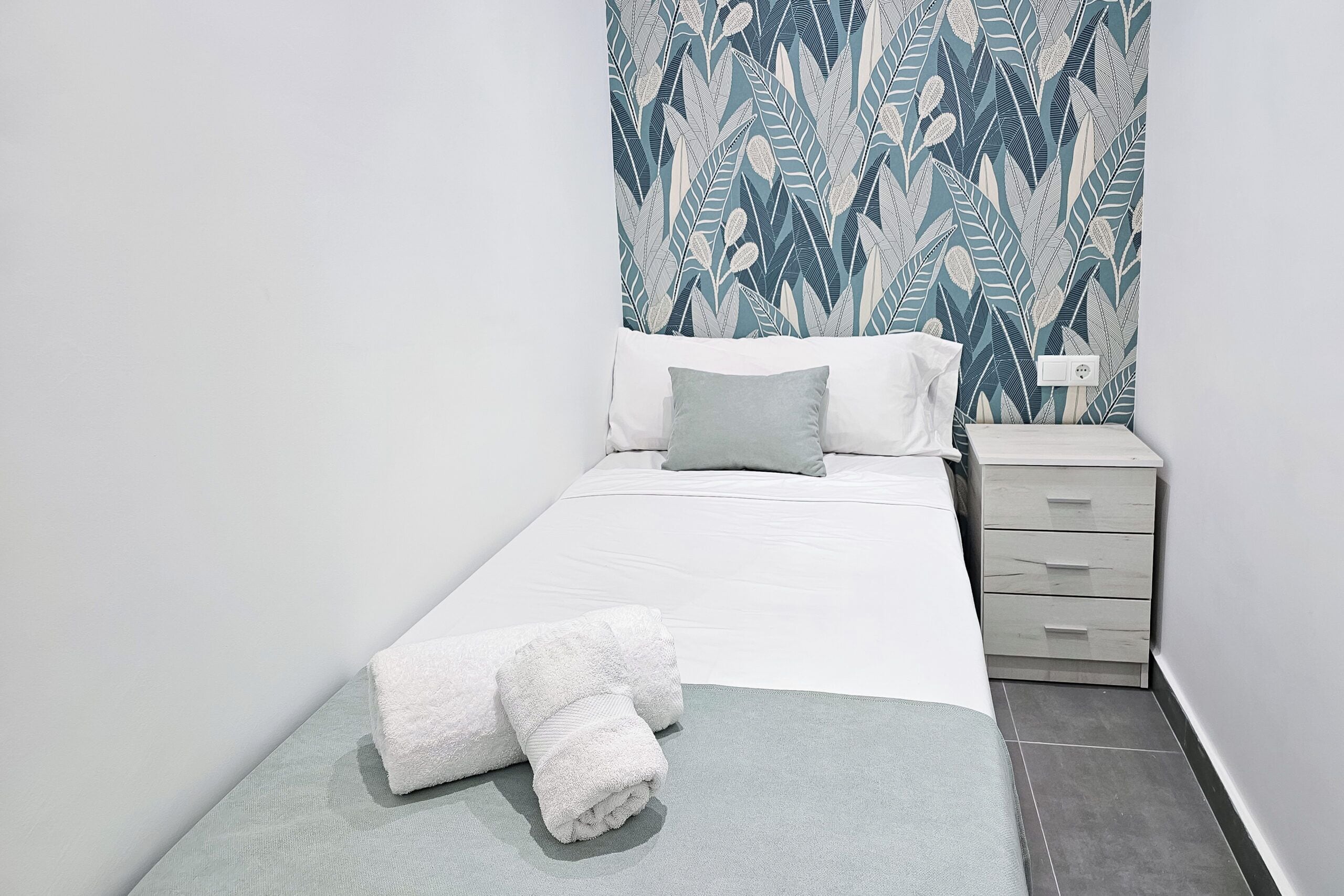 A neatly made bed with white linens and a folded towel, a bedside table with a lamp, and a patterned wall.