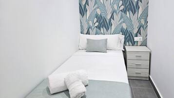 A neatly made bed with white linens and a folded towel, a bedside table with a lamp, and a patterned wall.