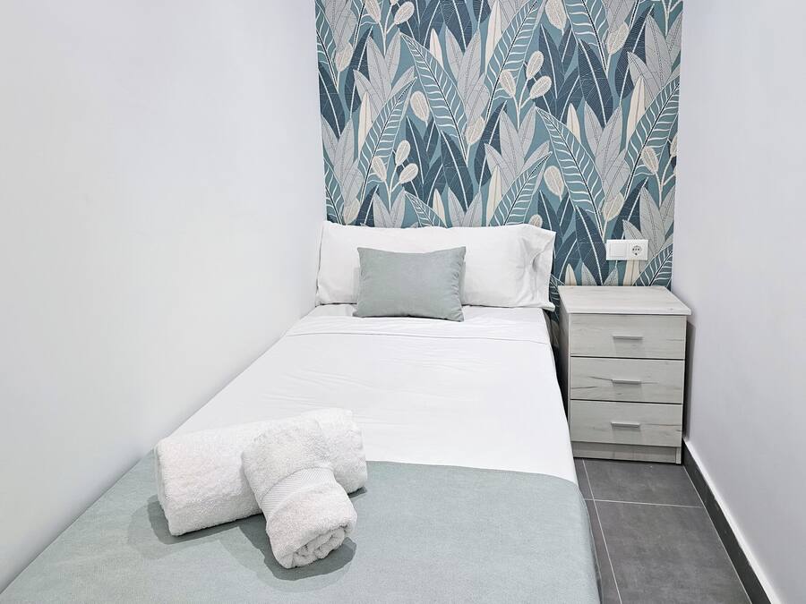 A neatly made bed with white linens and a folded towel, a bedside table with a lamp, and a patterned wall.