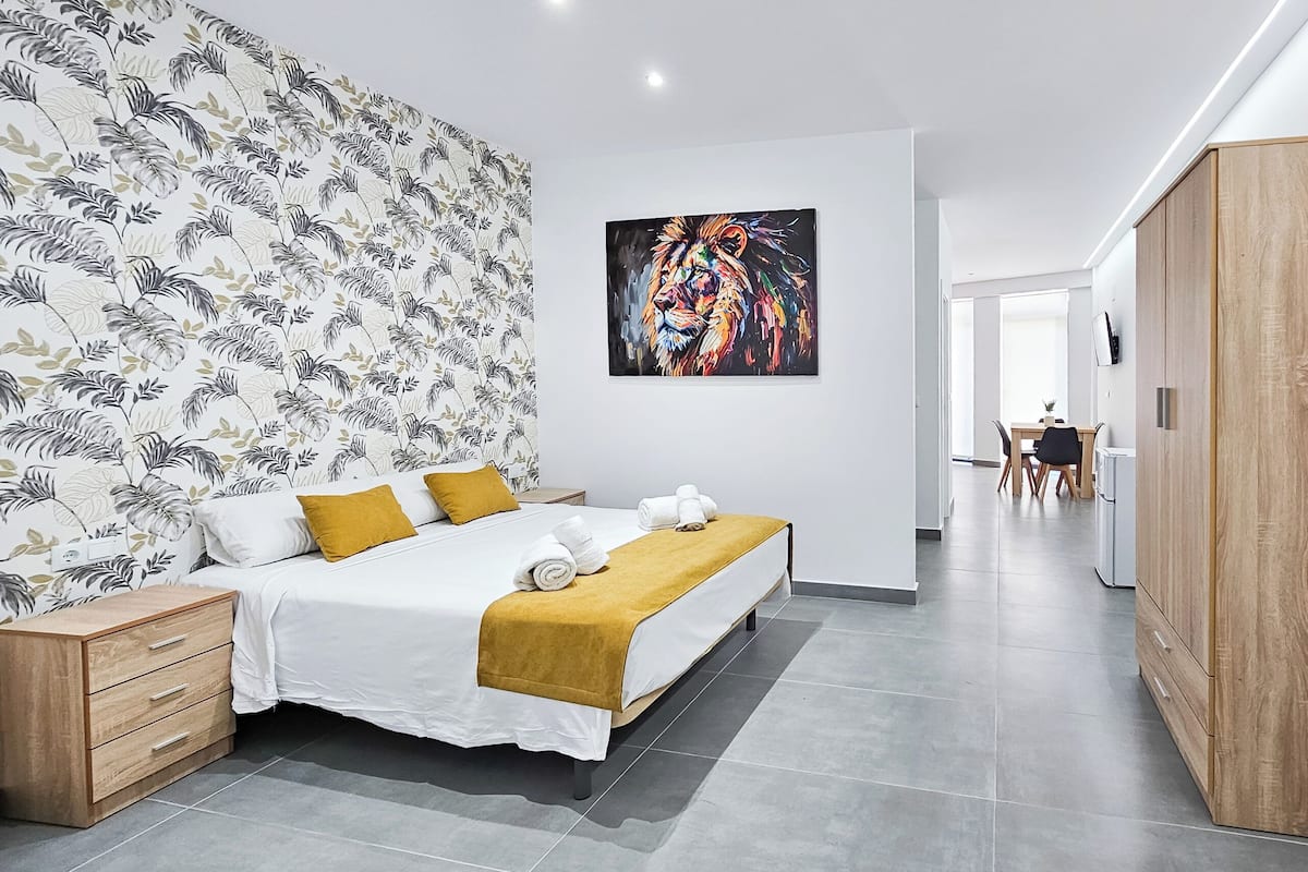 A modern bedroom with a large bed, a wooden wardrobe, a wall with a floral wallpaper, and a colorful painting.