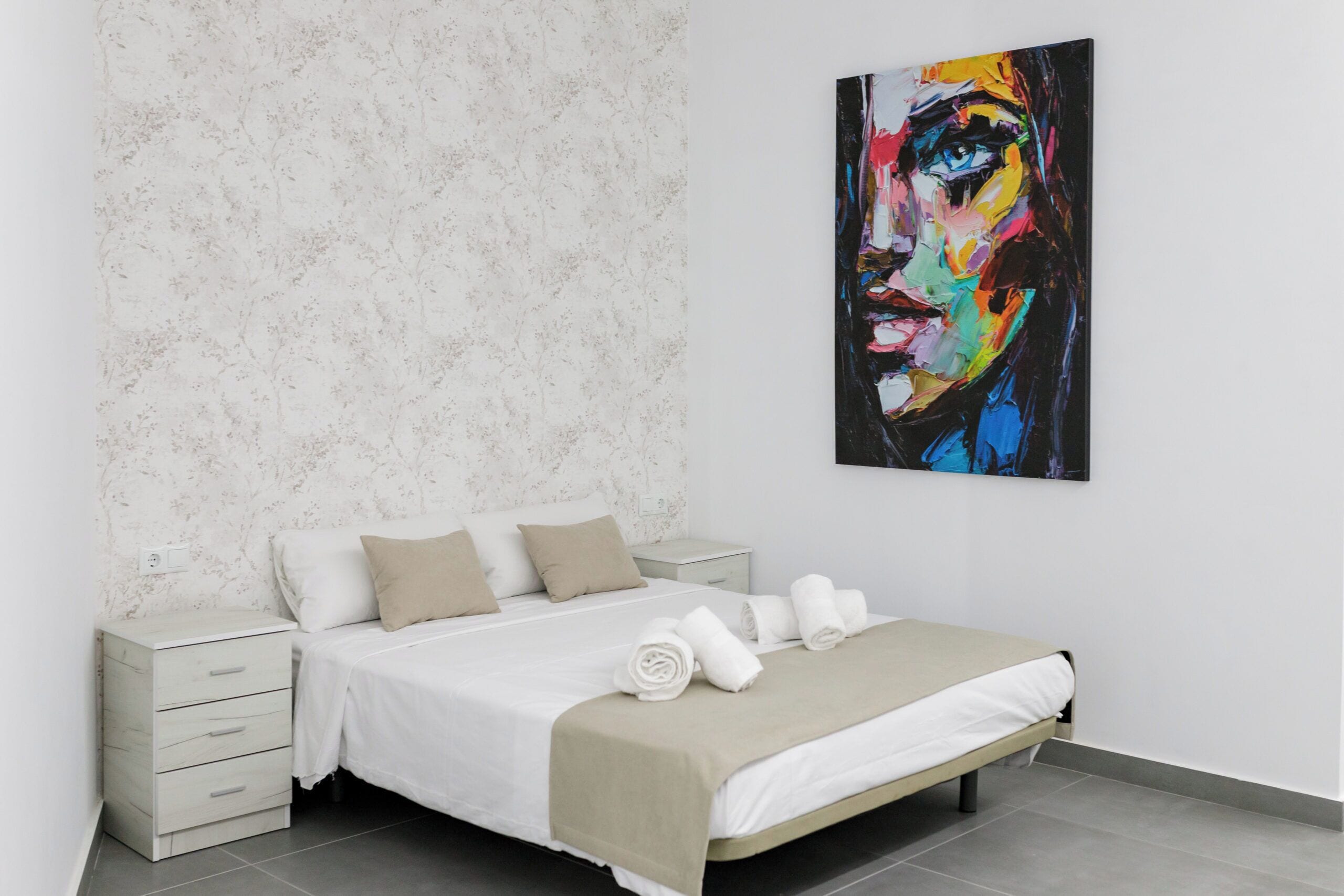 A modern bedroom with a bed, white nightstands, and a large abstract painting on the wall.