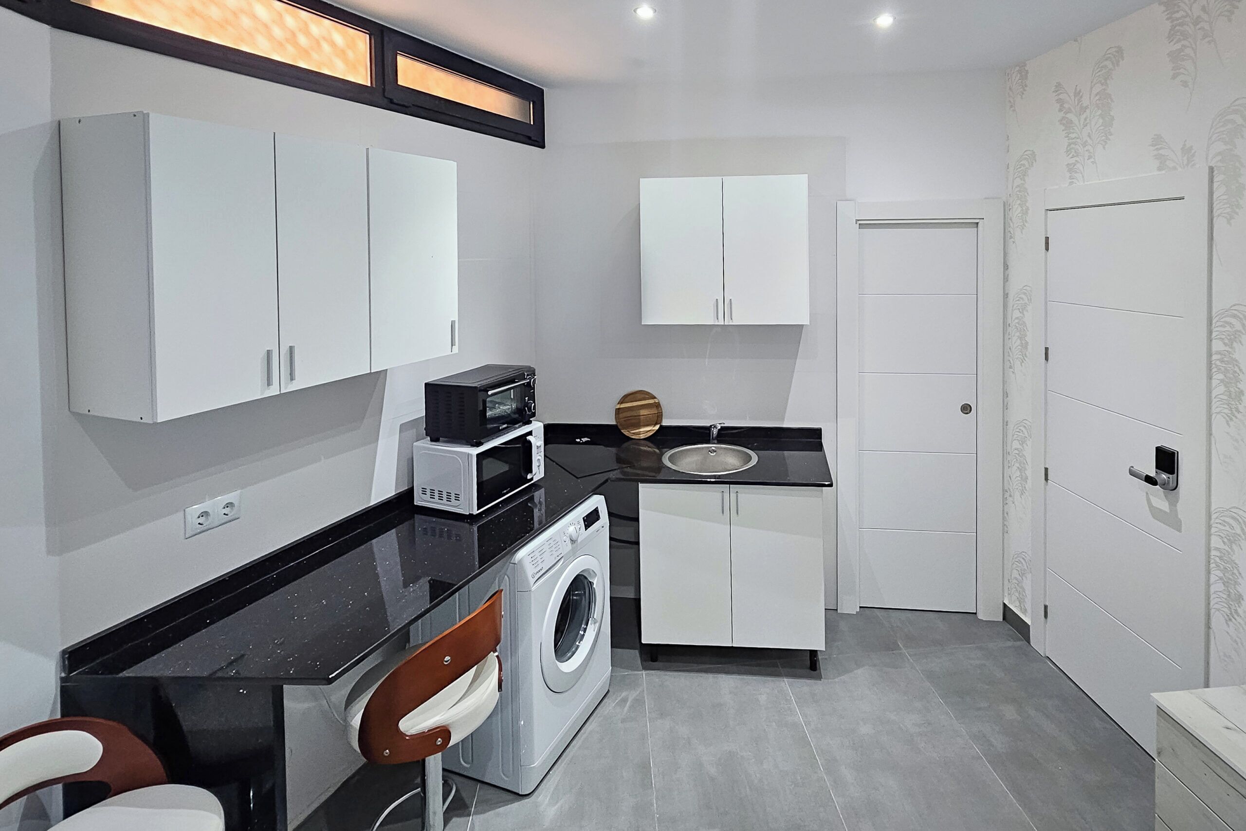 Une cuisine compacte avec des meubles blancs, un plan de travail noir, un four à micro-ondes, un évier et un lave-linge.