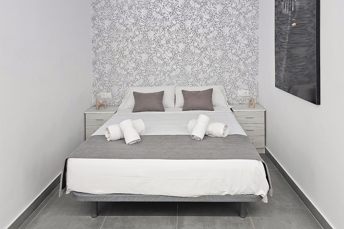 A neatly made bed with white and gray bedding, two pillows, and two rolled towels on the nightstand beside it.