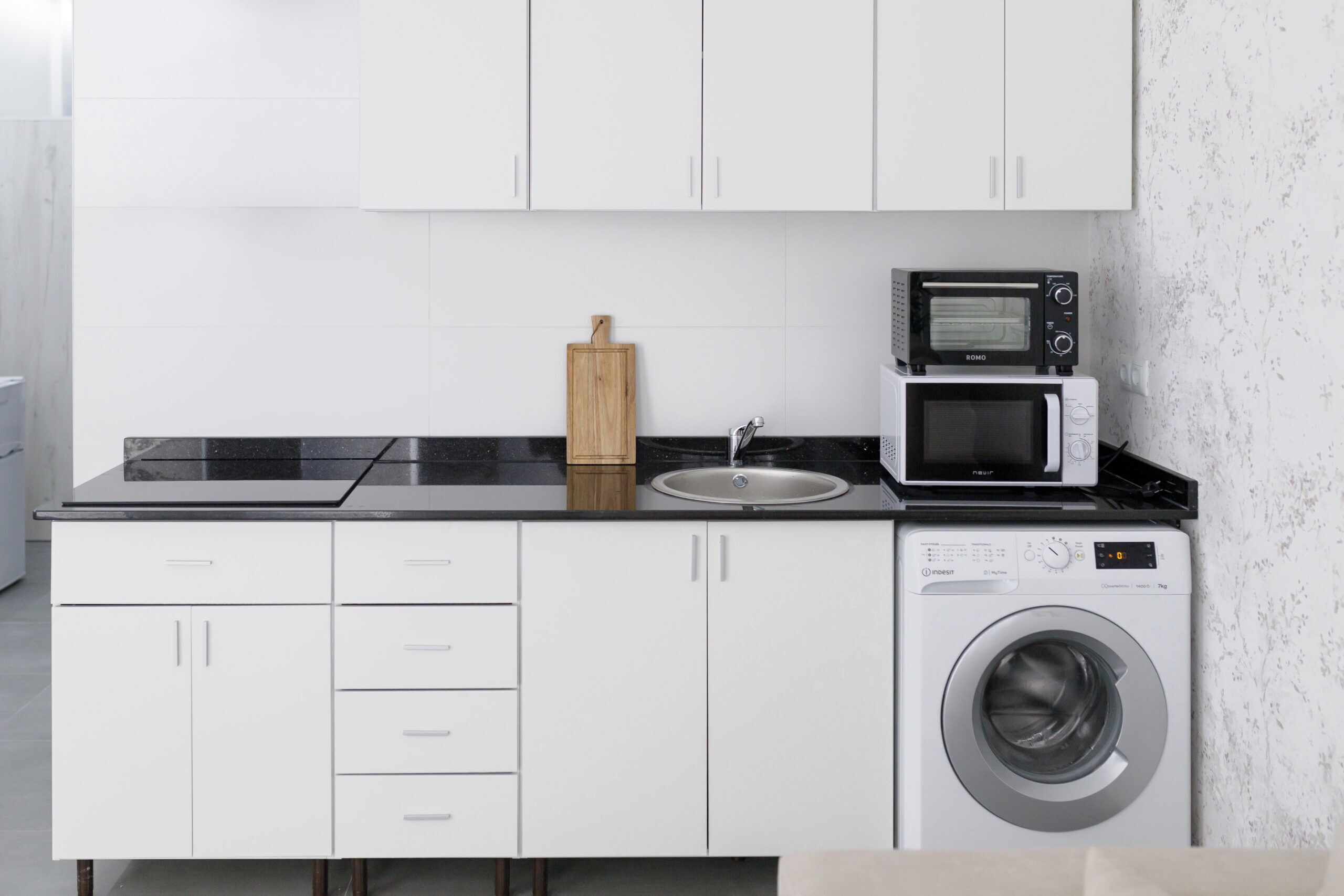 Cocina compacta con armarios blancos, encimera negra, microondas, tostadora y lavadora.