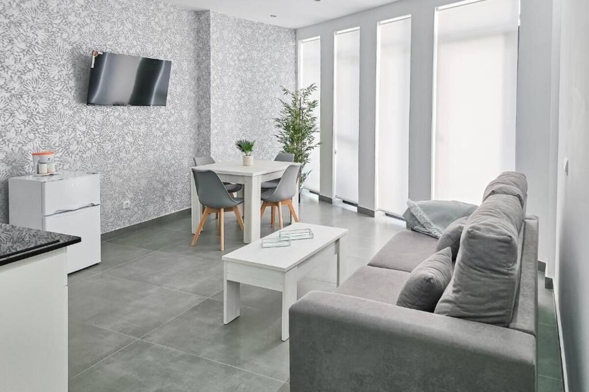 A modern kitchen with a dining area, a grey sofa, and a flat-screen TV mounted on the wall.