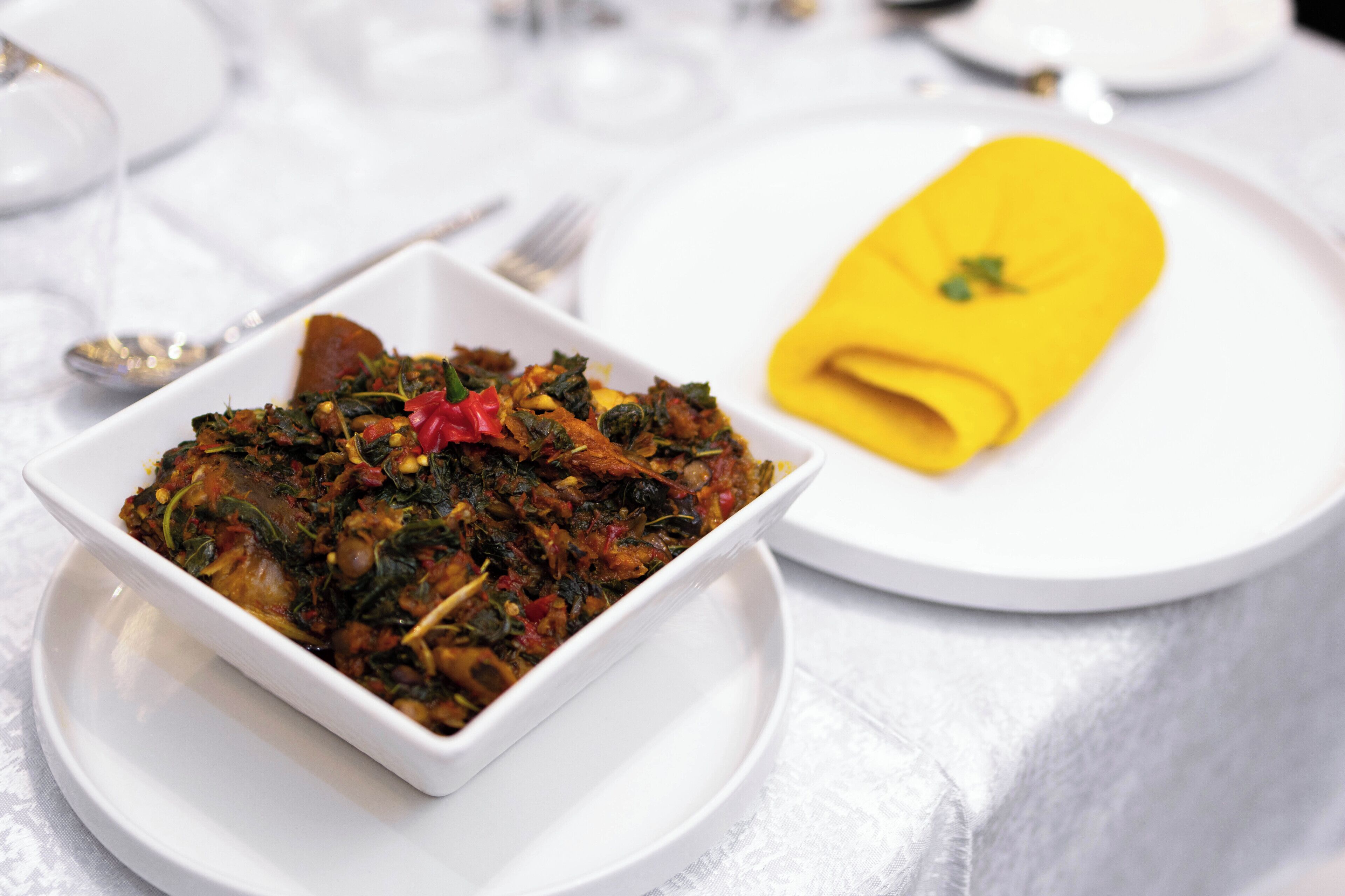A white rectangular dish with a colorful vegetable dish, a yellow napkin, and a white plate with a spoon and fork on a table with a white tablecloth.