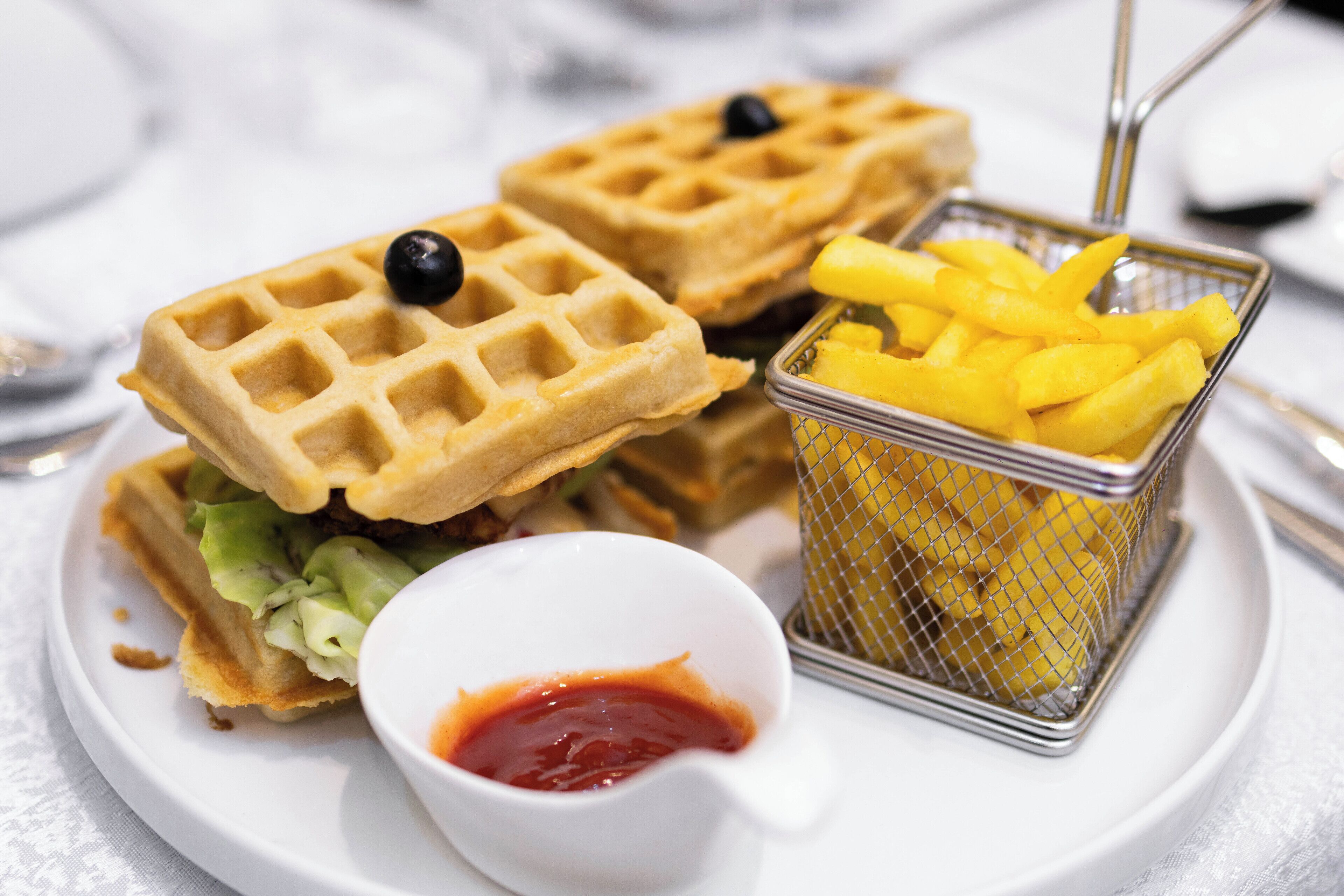 A plate with two sandwiches, a small bowl of ketchup, and a basket of fries.