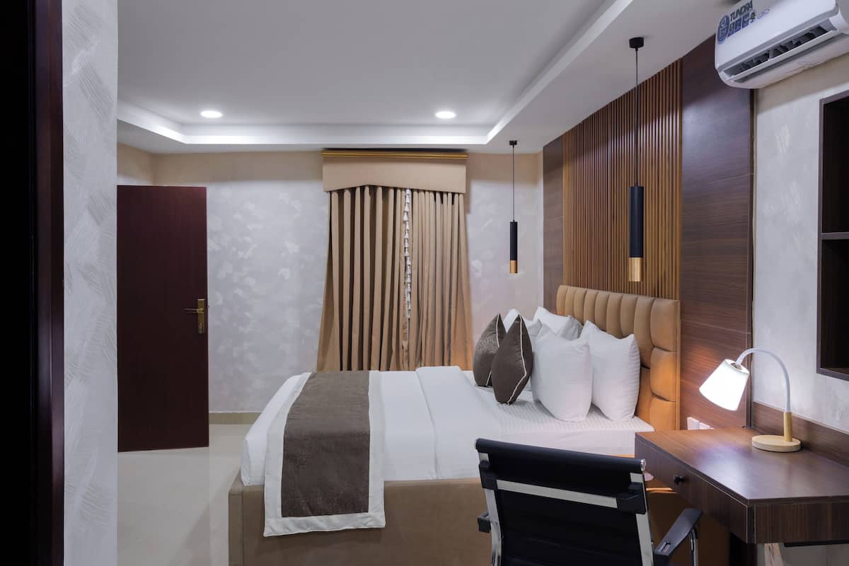 A hotel room with a bed, bedside table, lamp, and an air conditioning unit.