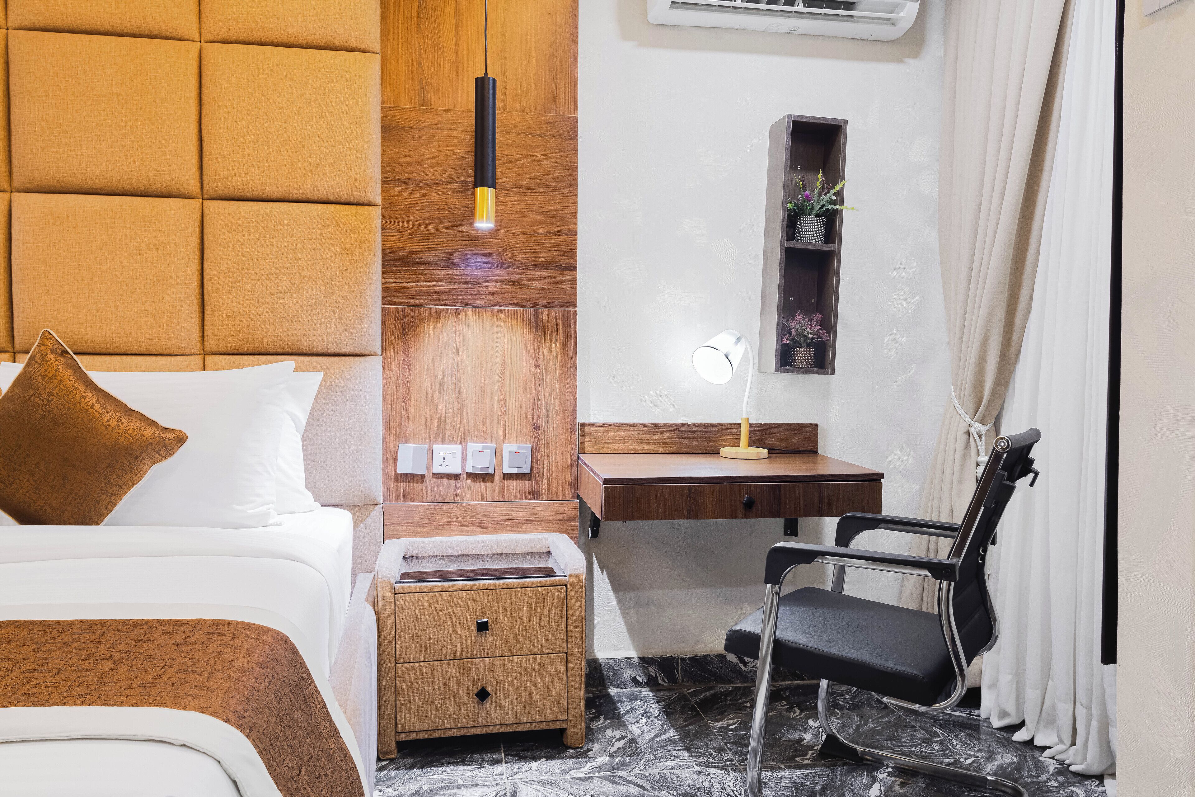 Habitación de hotel con cama, escritorio, silla, mesita de noche y estante con plantas.