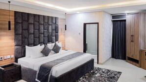 A modern bedroom with a large bed, a wooden headboard, a bedside table, a wardrobe, and a glass door leading to a bathroom. - BON Hotel Kabuwol (Ibadan)