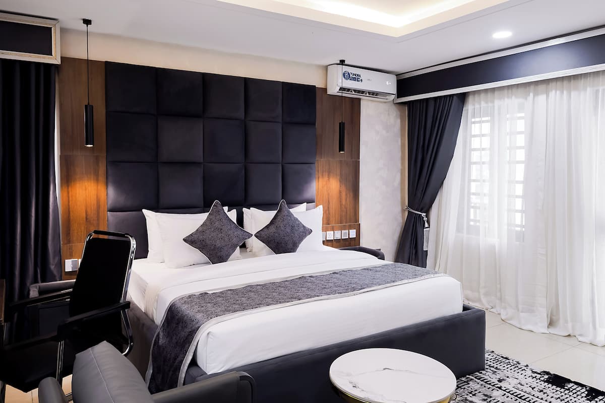 A modern hotel room with a large bed, a bedside table, a chair, a round coffee table, and a large window with curtains.