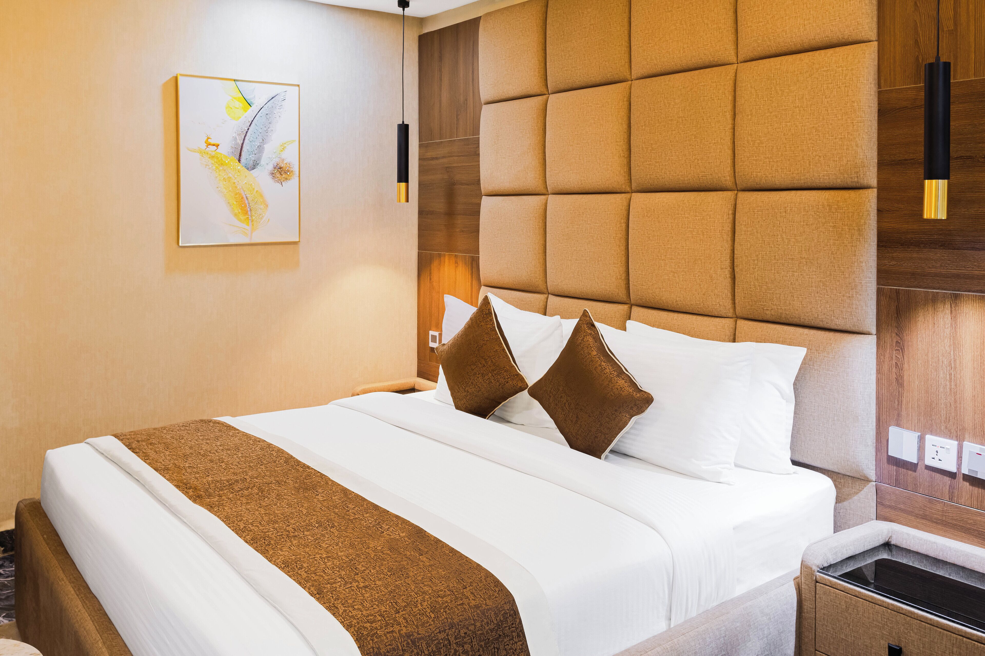 A hotel room with a large bed, a headboard with a wooden panel design, a bedside table with a lamp, and a painting on the wall.