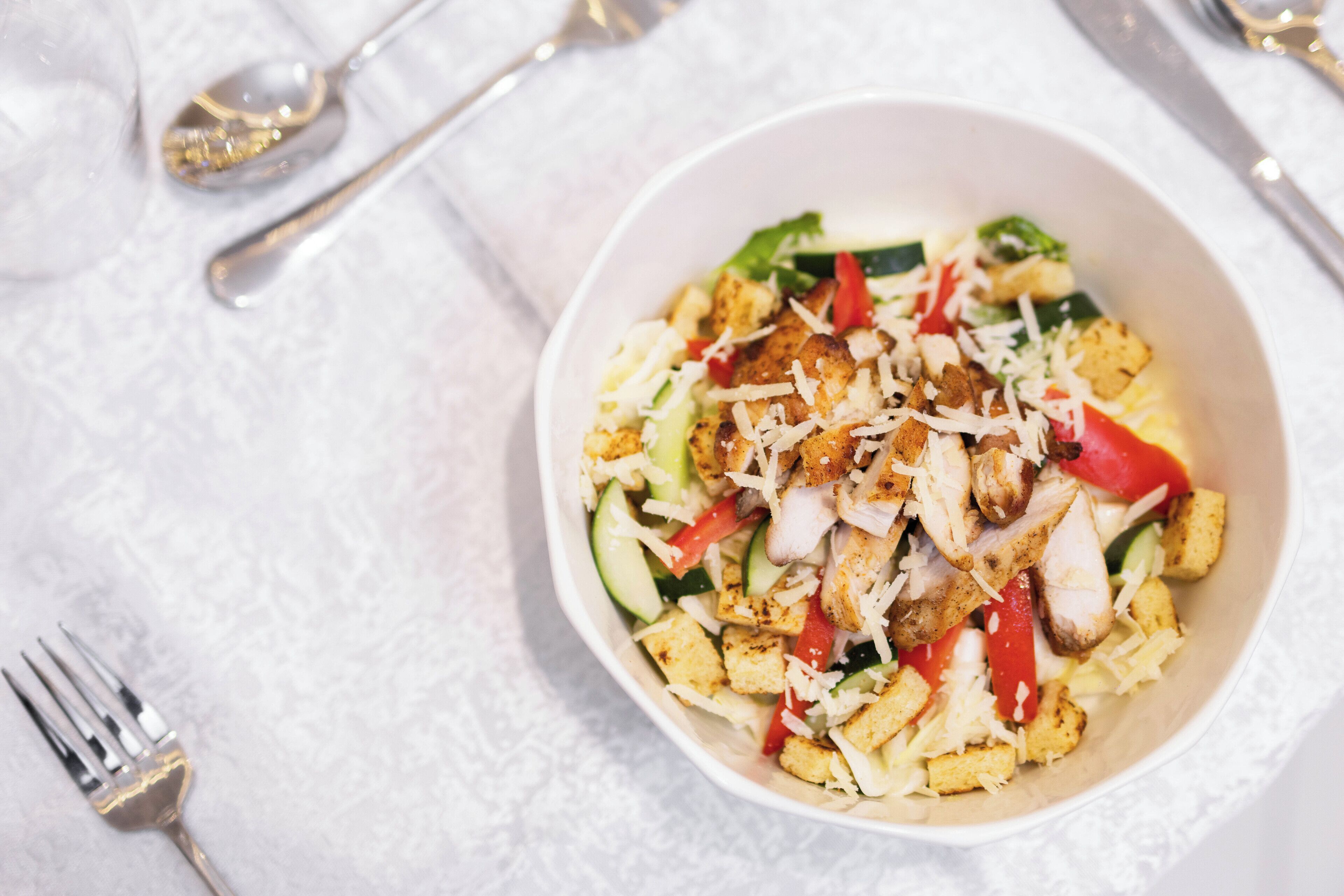 A white bowl of salad with croutons, shredded cheese, and red peppers on a white tablecloth.