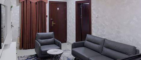 A modern living room with a grey sofa, a grey armchair, and a small round table. There are two wooden doors and a large window.