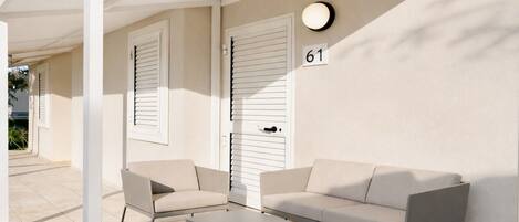 A covered outdoor seating area with two sofas and a table, a door with shutters, and a wall-mounted light fixture.