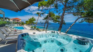 Un bain à remous avec une piscine et des chaises longues, donnant sur une étendue d’eau bordée d’arbres, avec un bâtiment visible au loin.