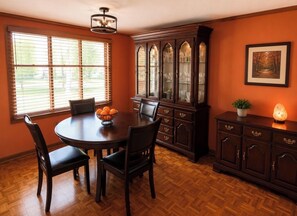 Un comedor con una mesa redonda, sillas, un mueble con puertas de vidrio y una ventana con persianas.