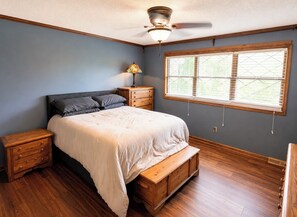 Un dormitorio con cama, mesitas de noche de madera, un banco, una ventana con persianas y un ventilador de techo.