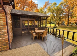 Un patio techado con una mesa y sillas de madera, un muro de ladrillo y vista a un parque con árboles.