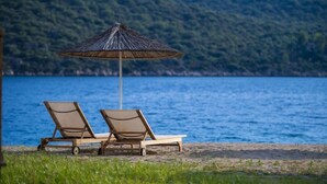 Two wooden lounge chairs with a thatched umbrella on a beach with a view of a calm sea and green hills in the background. - The Doria Yacht Club Kas Hotel (Kaş)