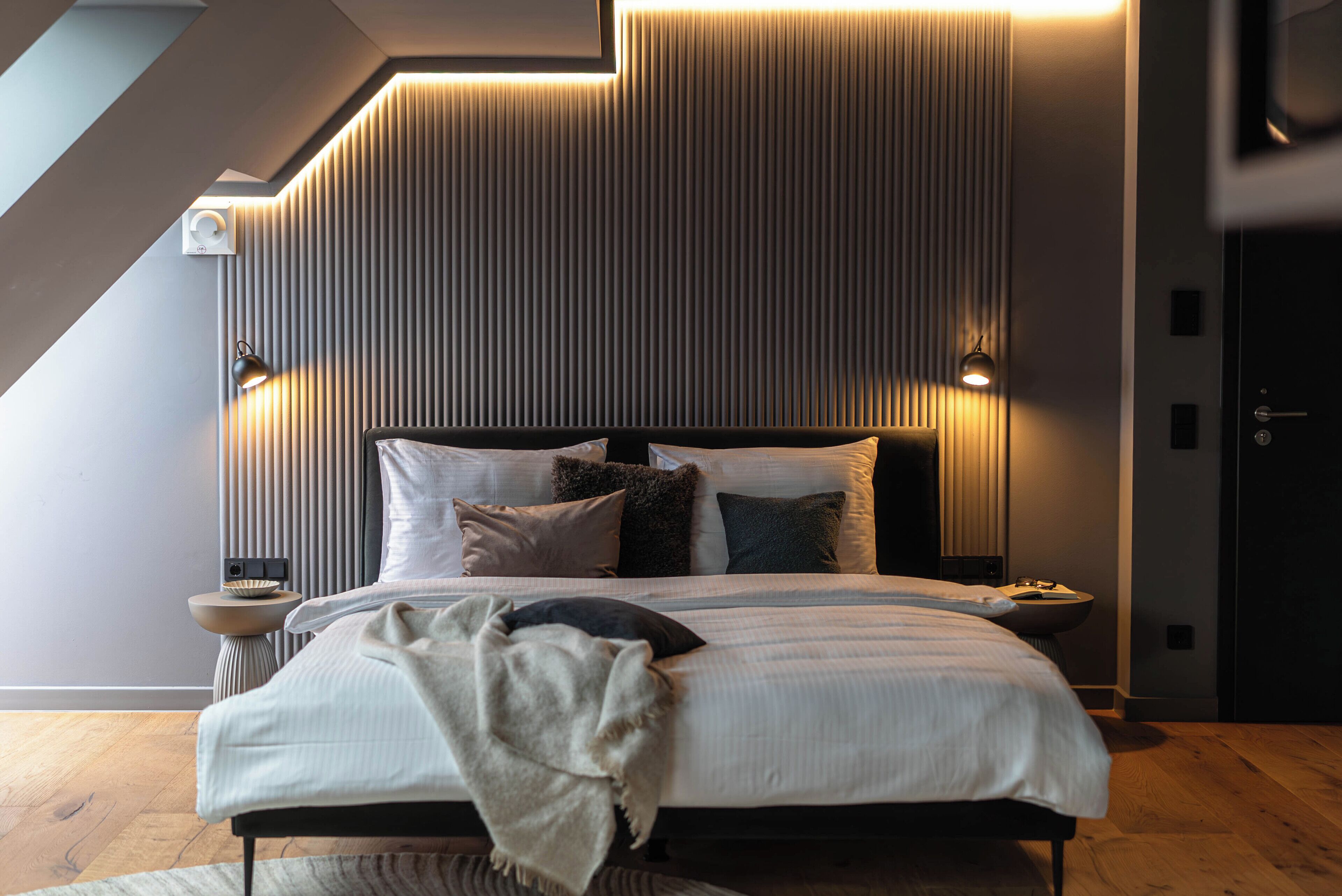 Een moderne slaapkamer met een schuine plafond, een groot bed met veel kussens, bedlampen en een klein tafeltje met een schaal.
