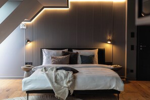 A modern bedroom with a sloped ceiling, a large bed with multiple pillows, bedside lamps, and a small table with a bowl. - Conlon House Central (Munich)