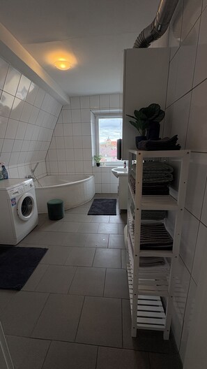 Une petite buanderie comprenant un lave-linge, un évier, une petite fenêtre et une étagère où sont pliées des serviettes.